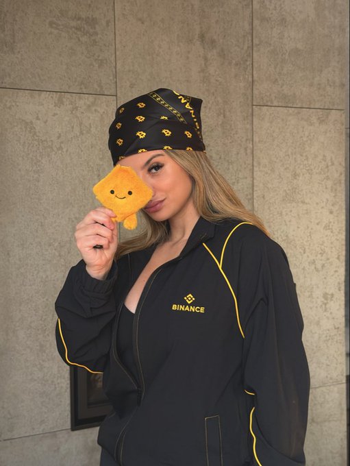 A woman poses in Binance-branded merchandise, wearing a black bandana and track jacket featuring the "BINANCE" logo in yellow. She holds up a smiling toast-shaped plush toy in front of one eye, playfully fitting the "GM" (Good Morning) crypto greeting vibe while promoting $BNB.