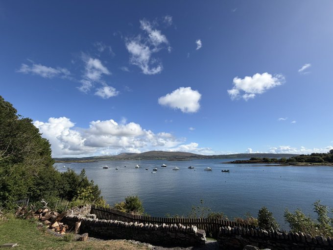 A scenic coastal view shows boats anchored in calm blue waters with rolling green hills in the distance under a bright sky dotted with white clouds, framed by lush foliage and a stone wall in the foreground.