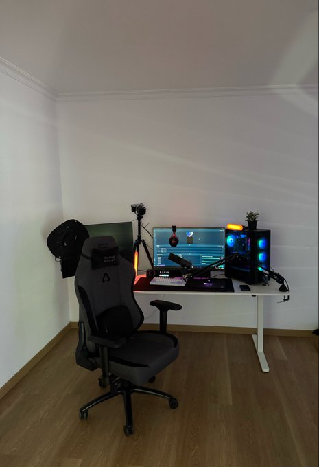 A gaming setup featuring a desktop with monitor displaying a video editing interface, RGB-lit PC tower, gaming chair, tripod, and a cap hanging on the wall, all arranged in a minimalist white room. The "GM chads Lock in" post reflects the focused, disciplined vibe of the clean, ready-to-work gaming/content creation space shown in the image.