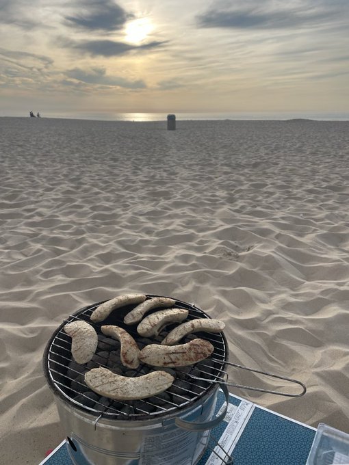 A serene beach scene at sunset featuring a portable charcoal grill cooking slices of flatbread or tortillas on the sand, with the ocean and a few distant figures visible in the background.