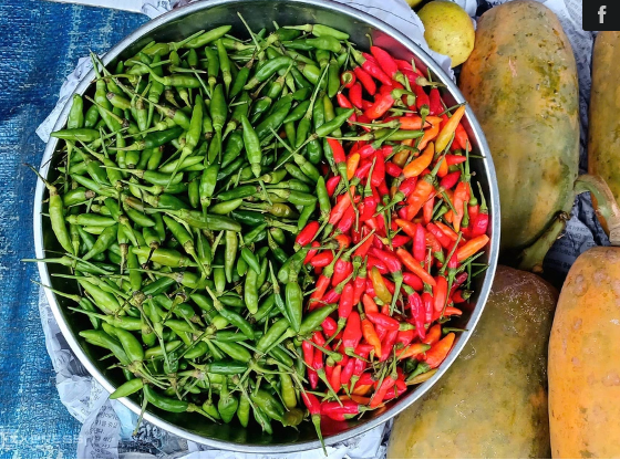 A metal bowl filled with green and red chili peppers, sitting next to papayas on newspaper.