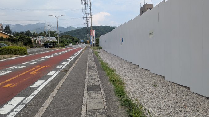 A street view shows a paved sidewalk and a red-painted bike lane next to a tall white construction barrier wall, with mountains and utility poles visible in the background.