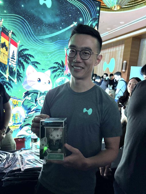 A smiling man wearing glasses holds up a "HYPFIRE" branded collectible figure in a box featuring a cat character, posing at what appears to be a branded event with vibrant neon artwork in the background—showcasing the sought-after merchandise the post encourages attendees to grab before they miss out.
