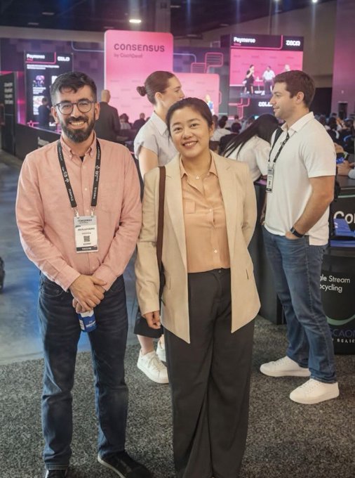 Two people, a man in a pink shirt and a woman in a tan blazer, pose together wearing lanyards at what appears to be the Consensus conference.