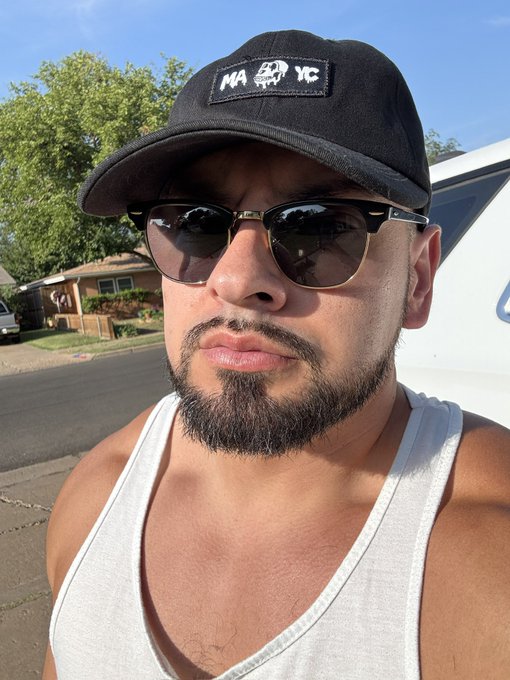 A man wearing sunglasses and a black cap with a "MAYC" patch featuring a skull design takes a selfie in a suburban street, visually echoing the post's "Born in the MAYC trenches" caption by displaying his affiliation with the Mutant Ape Yacht Club.