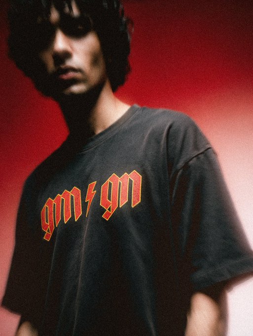 A young man with dark curly hair wears a black t-shirt with red gothic-style text, posing against a red gradient background.