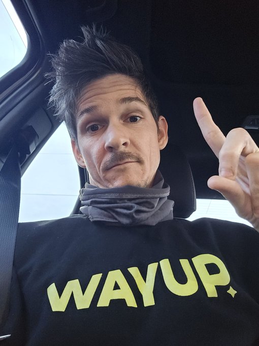 A man with a mustache sits in a car wearing a black 'WAYUP.' sweatshirt and a gray neck gaiter, making a hand gesture.
