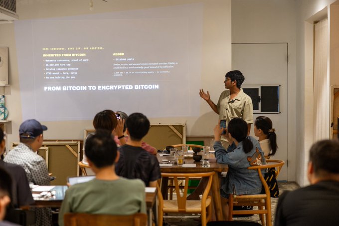 A presenter stands before a projection displaying "FROM BITCOIN TO ENCRYPTED BITCOIN" at a cryptocurrency meetup, gesturing toward an engaged audience in Taipei that is taking photos and listening attentively.