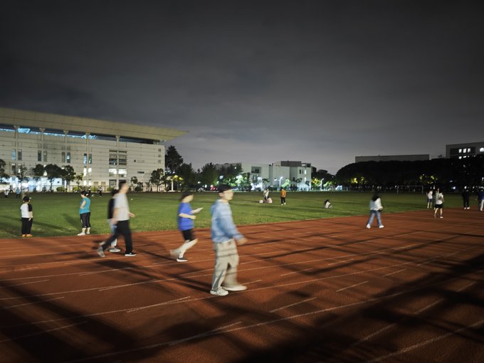Several people are walking and jogging on a red running track at night, with a large illuminated building visible in the background.
