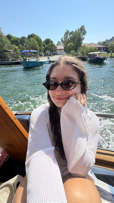 A selfie of a young woman wearing sunglasses and a white knit sweater on a boat, with turquoise water and other boats in the background.