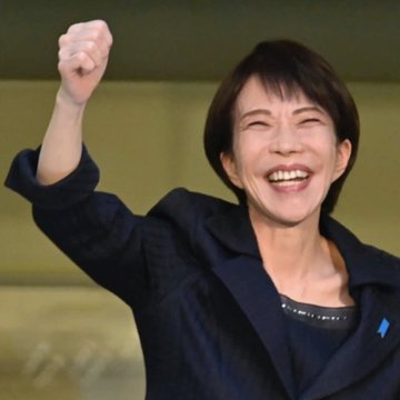 The image features a woman with short hair, wearing a black jacket, who appears to be smiling and waving her hand up in the air. The background of the photo shows a building, suggesting that she might be at an event or gathering related to Algorithand Japan. The caption on the post reads "JAPAN LOVES ALGORAND $ALGO @AlgorandJapan". This indicates that the woman is promoting a product or service associated with Algorithand Japan and possibly expressing her enthusiasm for it.