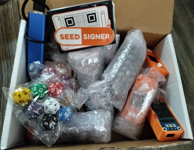 An open box containing a SEED SIGNER hardware wallet kit, with a sticker on top and bubble-wrapped components including colorful polyhedral dice and orange electronic devices inside.