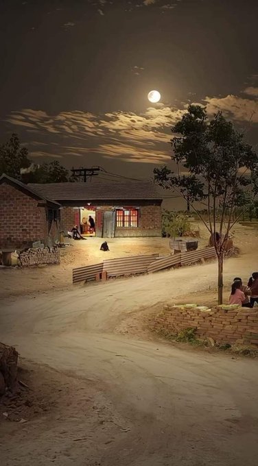 A nighttime rural scene showing a brick house illuminated under a bright full moon, with people gathered near the doorway and sitting by a dirt road.
