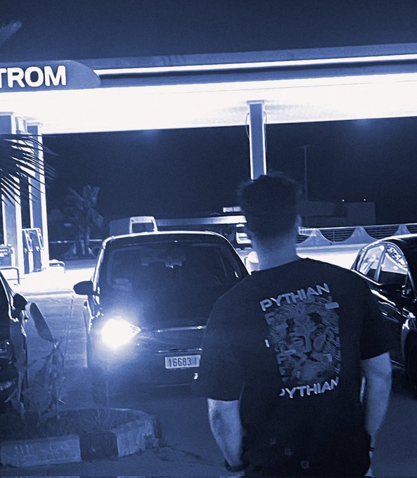 A nighttime photo of a person wearing a black 'PYTHIAN' t-shirt standing at a gas station, with a car visible in the background.