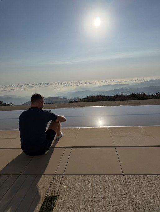 A man sits on a stone patio overlooking an infinity pool and a sea of clouds beneath a bright sun, gazing toward a horizon often nicknamed "above the heavens." The serene scene visually echoes the "TLI in the heavens" caption, suggesting tranquility and a heavenly view from a high vantage point.