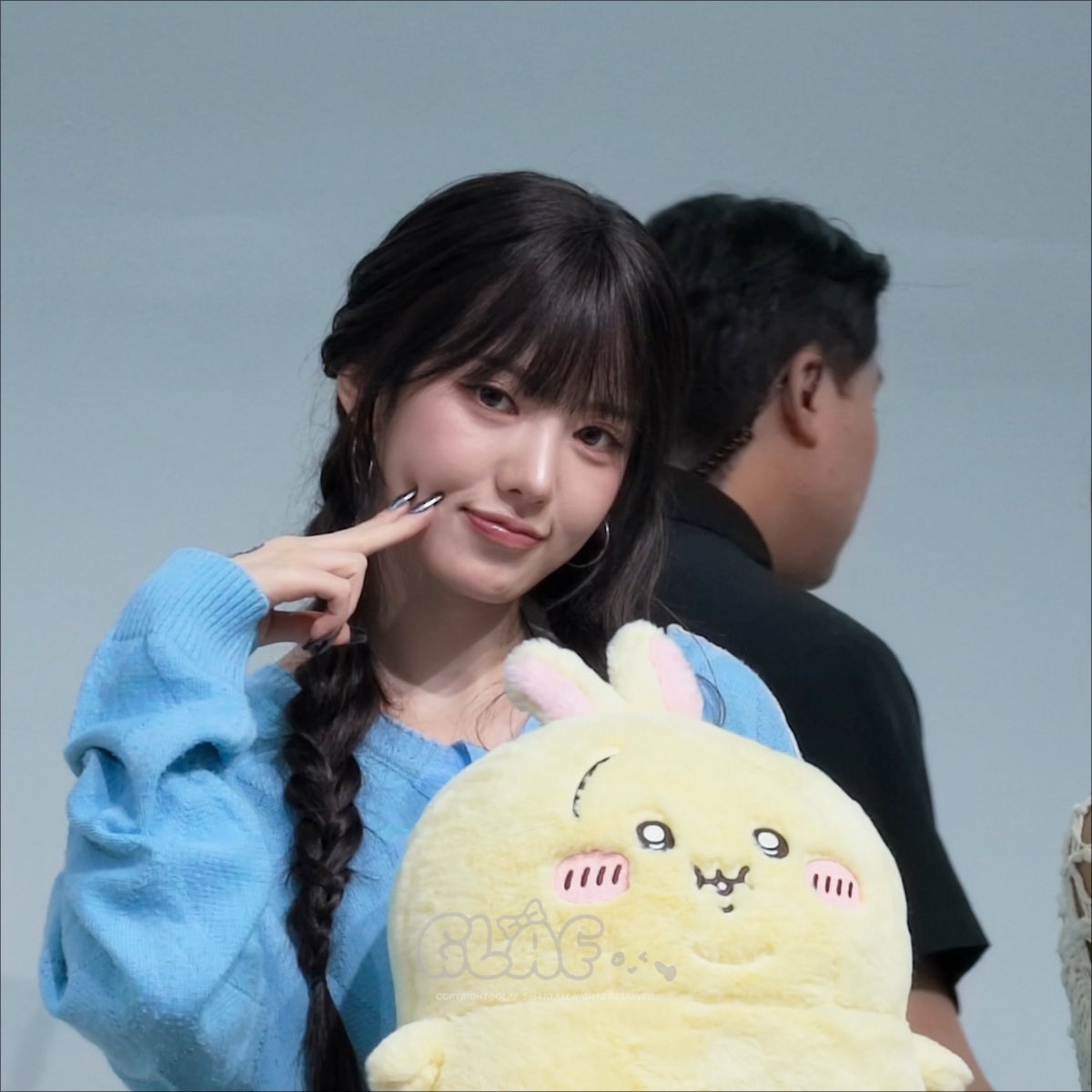 A young woman with dark braided hair wearing a blue sweater, smiling and pointing at her cheek while holding a yellow plush toy with bunny ears.