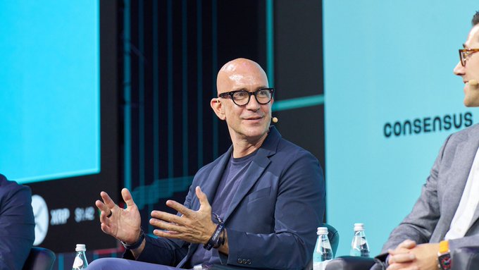 Satya Nadella speaks on stage at a Consensus event, gesturing with his hands while seated next to another panelist.
