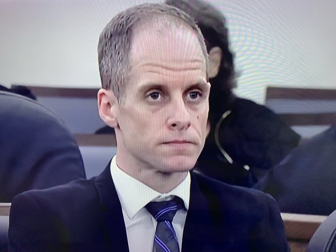 A man with short hair in a dark suit and striped tie looks on with a serious expression in what appears to be a courtroom setting.
