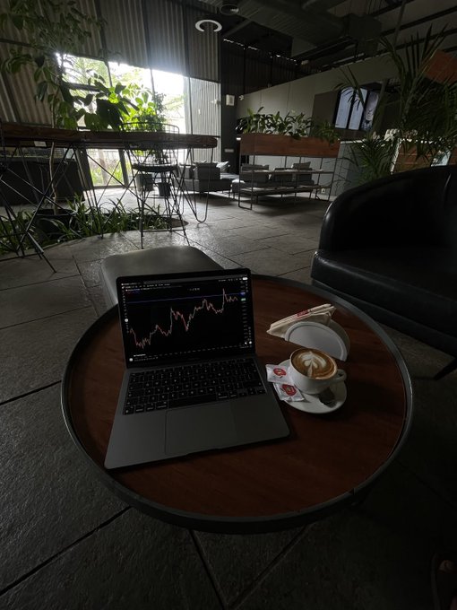 A laptop displaying a stock market chart sits on a round wooden table in a cafe, next to a cup of coffee with latte art.