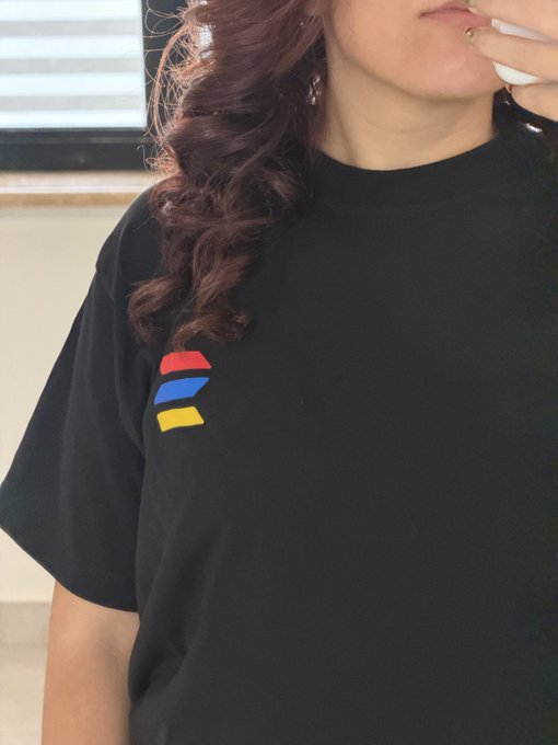 A close-up photo of a person with long reddish-brown hair wearing a black t-shirt with a small red, blue, and yellow striped logo on the chest, while holding a tissue to their mouth.