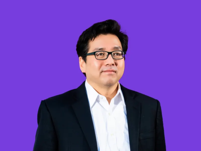Tom Lee, a well-known market strategist, stands against a purple background in a black suit and glasses. The image accompanies a post about his prediction that crypto, particularly ETH, could be the biggest FOMO trade if the Fed doesn't hike rates in September.