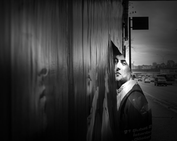 A black and white photograph of a man peeking around the corner of a large metal container, wearing a jacket with '21 Subst 2 Istanbul' text, set against an urban backdrop with cars and overpasses.