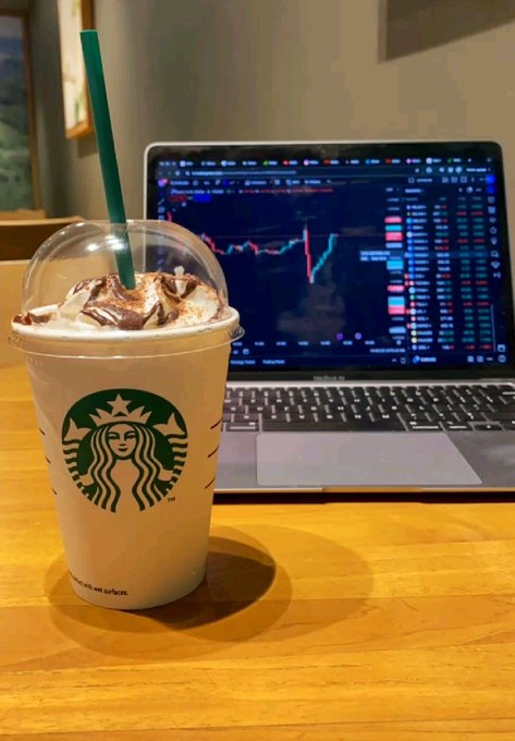A Starbucks Frappuccino topped with whipped cream, chocolate drizzle, and a green straw sits on a wooden table in front of a laptop displaying a trading chart.
