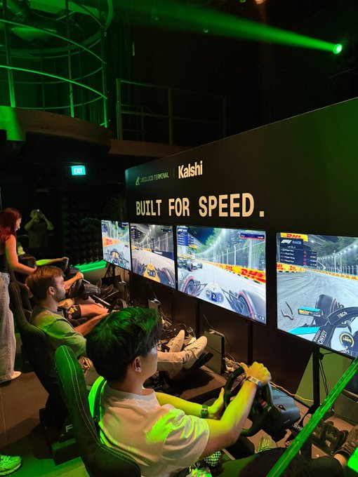 Two people playing a racing simulator game in a room lit with green light, with the text 'Kalshi BUILT FOR SPEED.' displayed on the wall behind the monitors.