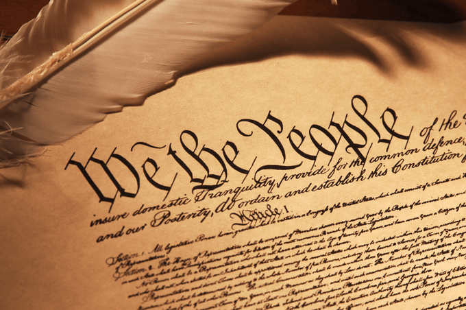 A close-up, sepia-toned view of the U.S. Constitution's opening words "We the People of the" written in elegant calligraphy, with a quill pen resting in the upper left corner. The image directly ties to ConstitutionDAO's $PEOPLE token, as the famous "We the People" phrase inspired the DAO's mission to crowdfund a community purchase of this historic document.