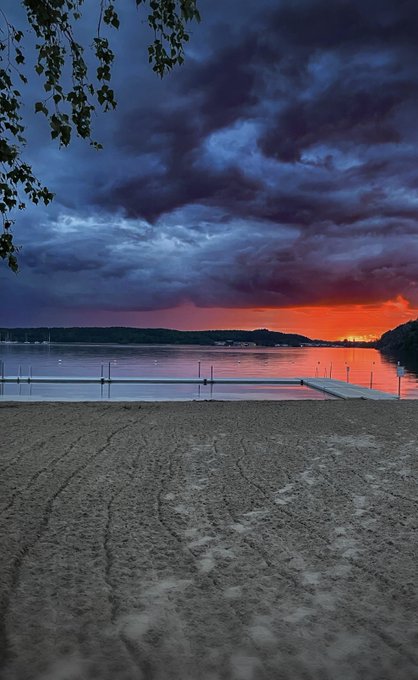 A serene beach scene at sunset with dramatic dark clouds overhead and orange light reflecting on the calm water, featuring a wooden dock and footprints in the sand.