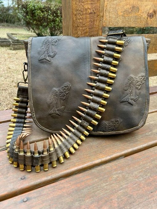 A distressed brown leather messenger bag adorned with embossed designs of pistons and hot rod cars, displayed on a wooden bench alongside an ammo belt draped with brass-cased cartridges.