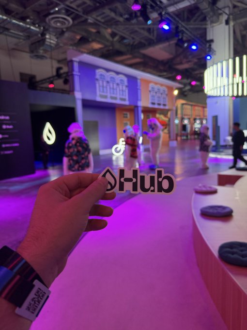 A photo taken at a tech conference showing a hand holding a 'Hub' sign/merchandise, with colorful neon-lit stage installations and attendees visible in the blurred background.