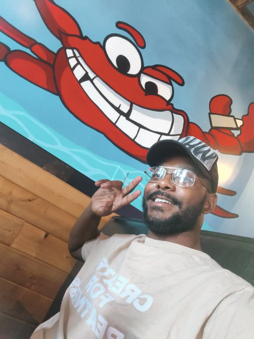 A man wearing glasses and a 'Daddy' cap poses with a hand gesture in front of a large cartoon red lobster mural painted on a blue wall.