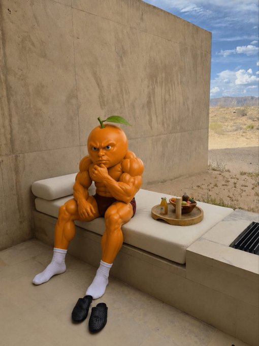 A muscular anthropomorphic orange with a furrowed brow sits pensively on a stone bench in a desert setting, beside a tray of fruits and accompanied by a tray of drinks, with white socks and black slippers on his feet—visually echoing the "roller coaster ride" of emotions tied to trading $Udin.