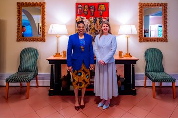 The image shows two women standing next to each other in a room with chairs around them. One woman is wearing a blue jacket, while the other has a white dress. They are both smiling at the camera. The room features a large mirror on one wall and several lamps placed throughout the space. A painting can be seen hanging above the table where they stand.
