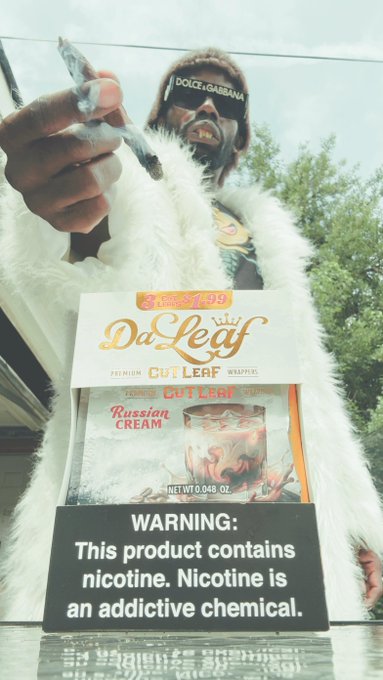 A person in a furry outfit wearing Dolce & Gabbana sunglasses holds a package of DaLeaf 'Russian Cream' Cut Leaf, with a prominent warning about nicotine being an addictive chemical.
