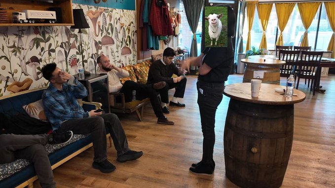 Photo of several young men relaxing in a lounge/cafe setting with patterned wallpaper, wooden barrel tables, and couches, with one person standing in the foreground.