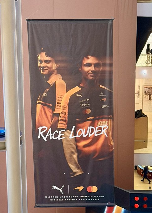 A large banner for the McLaren Mastercard Formula 1 Team displaying two drivers in racing suits with sponsor logos (Dell, OKX, Puma) and the slogan 'RACE LOUDER,' hanging in what appears to be a mall or public space.