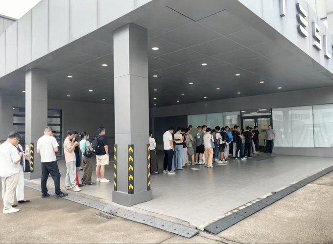 A crowd of people waits in line outside a building with partial signage reading "SE" visible on the facade, suggesting a queue at a showroom or service center that aligns with the caption "Tesla Lines are back on the Menu!!" indicating increased demand or interest in Tesla vehicles or services.