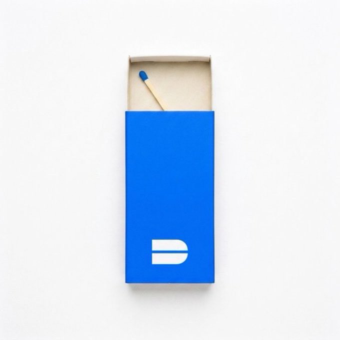 An open blue matchbox on a white background showing a single wooden match with a blue tip, featuring a white stylized "D" logo on the front.