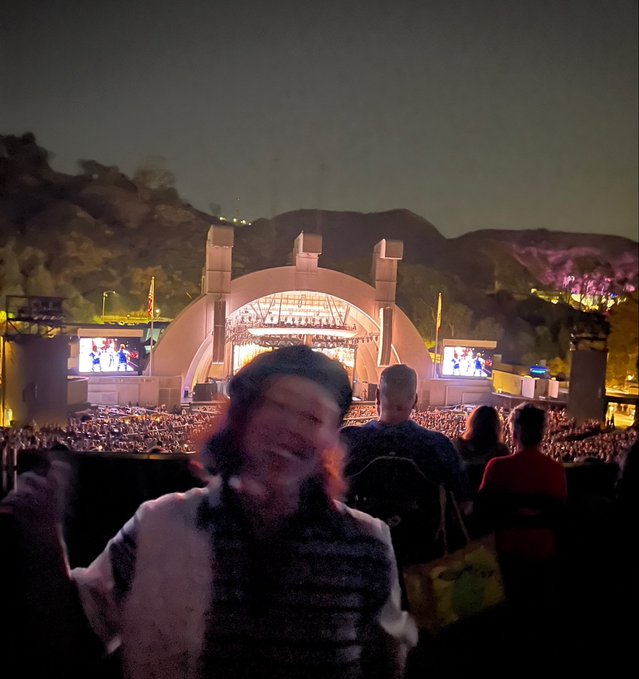 A blurry nighttime photo of a smiling person in a beanie at an outdoor concert venue, with the iconic Hollywood Bowl stage and mountains visible in the background.
