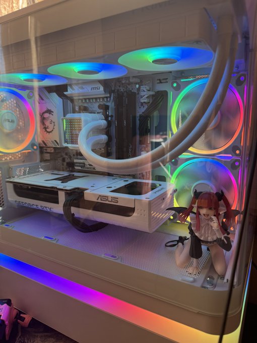 A custom-built gaming PC inside a white case with colorful RGB lighting on the fans, featuring MSI and ASUS components, liquid cooling tubes, and a small anime figurine decoration on the bottom.