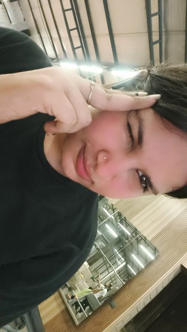 A close-up selfie of a young woman with dark hair smiling and giving a thumbs up near her face, taken indoors with a ceiling visible in the background.