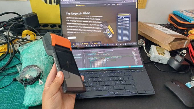 A hand holds an orange and black hardware wallet device in front of a laptop displaying an article titled "The Dogecoin Wallet" with a code editor open below it.