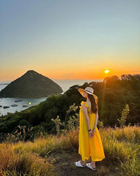 A woman in a yellow dress and wide-brimmed hat stands on a grassy hill overlooking a tropical bay with a conical island at sunset.