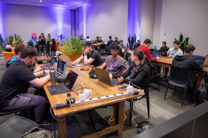 The image shows a group of people sitting around tables, each with their laptop open and focused on their work or conversation. The room has a large window that allows natural light to enter, creating a comfortable atmosphere for productivity. There are several potted plants placed throughout the space, adding a touch of greenery and life to the environment. A person is wearing a backpack in the foreground, possibly indicating they have brought some personal items with them while working or socializing at this event. The image also contains text that reads "overheard at ethconf: ?" which suggests that someone might be discussing or sharing information about an event happening at the conference center where people are gathered to work on their laptops.