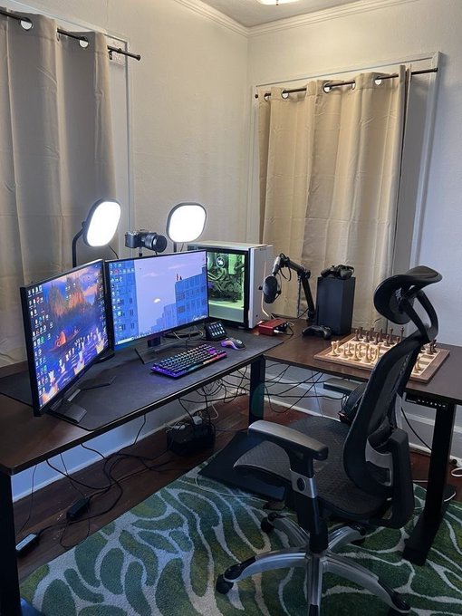 A home computer desk setup featuring a triple-monitor display, RGB keyboard, gaming chair, and panel lighting, with a chess board placed on the right side of the L-shaped desk.
