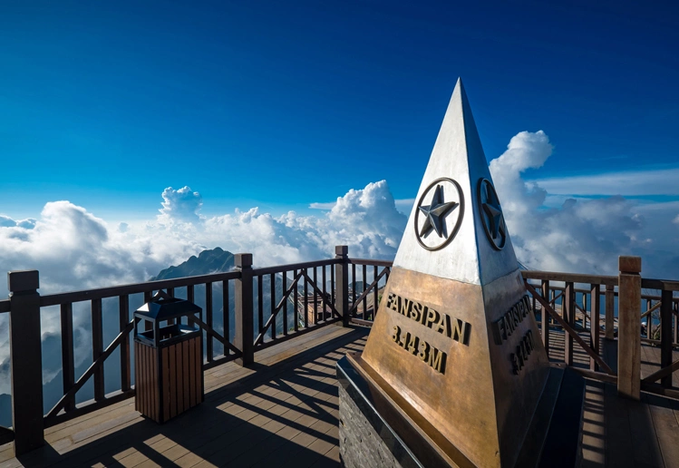 A stone obelisk marker on a wooden observation deck above the clouds at Fansipan, with mountains visible in the distance under a bright blue sky.