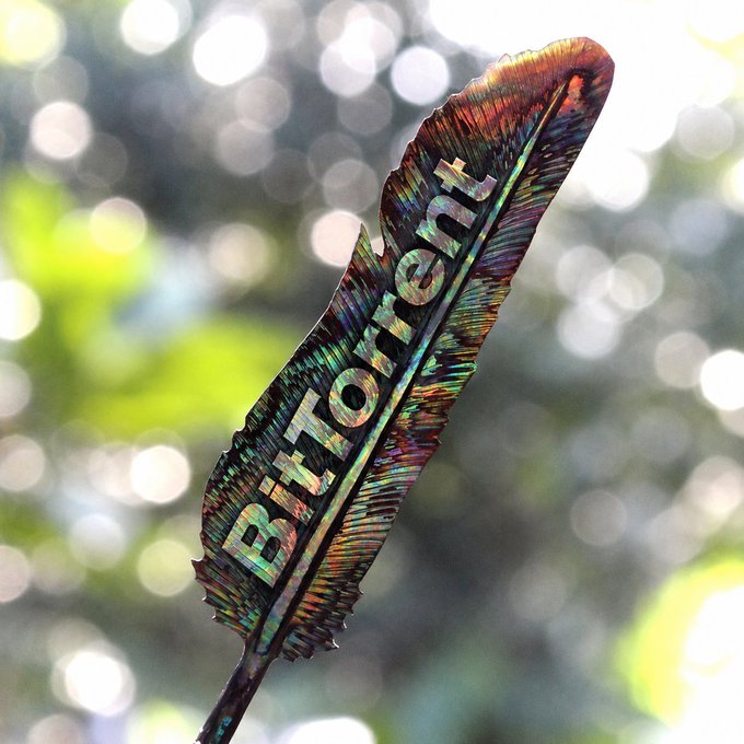 A iridescent feather displays the word "Breton" cut through its center, set against a softly blurred background of bokeh light. The feather's rainbow sheen and upward sweep visually echo the post's motivational message of letting creative ideas flow.