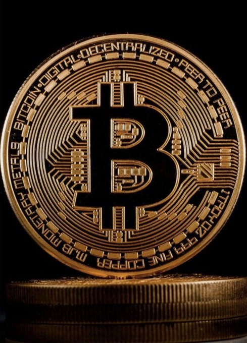 A close-up photograph of a gold Bitcoin token standing upright against a black background, with the text 'BITCOIN DIGITAL DECENTRALIZED PEER TO PEER' and 'MJB MONETARY METALS FINE COPPER 999 FINE ZINC 999 FINE' engraved around its edges.